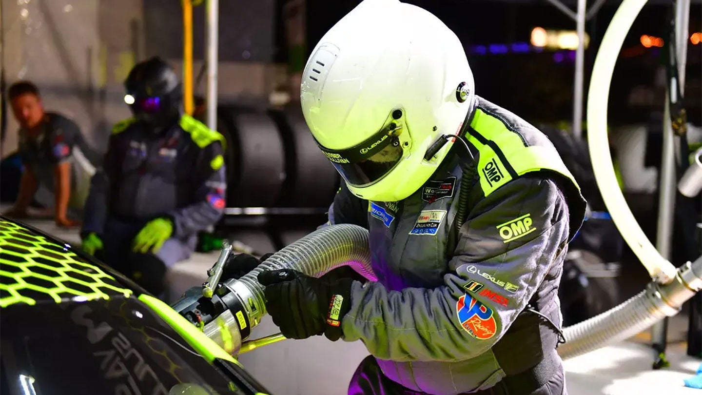 Pit crew member in a white helmet and gray racing suit refueling a car during a night pit stop.